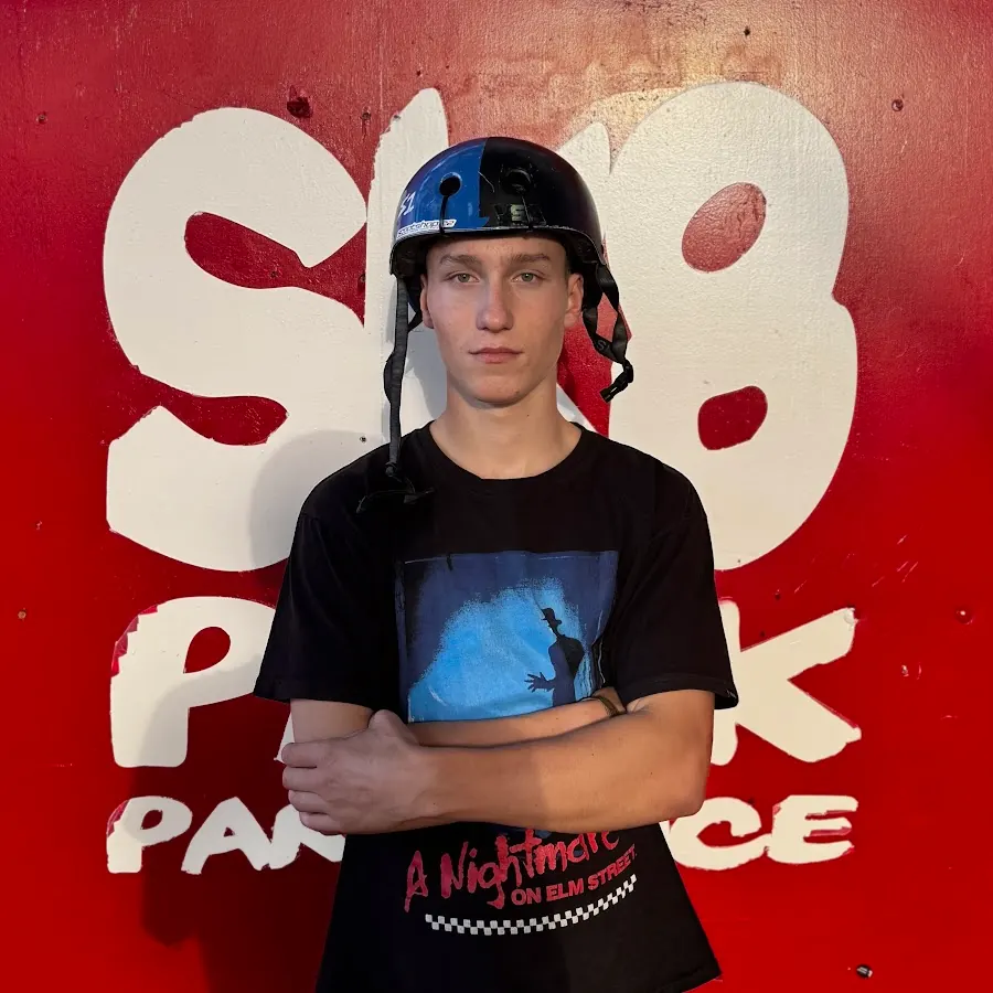 Saša Vaňous se založenýma rukama a helmou před červenou zdí SK8 Parku Pardubice